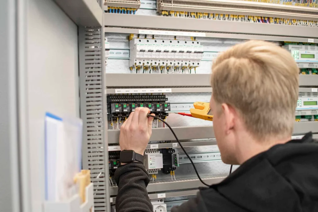 Tekniker med mätinstrument installerar IMD-mätning i en bostadsrättsförenings elcentral, med kontaktorer, automatsäkringar och mätinsamlingsenheter synliga i skåpet