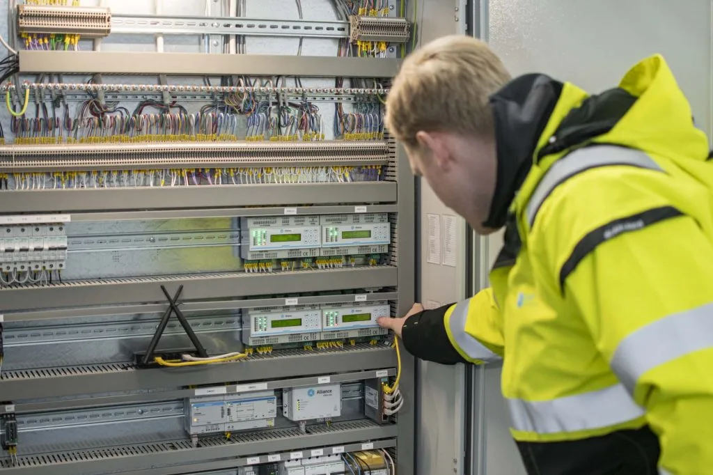 Tekniker i gul arbetsjacka utför IMD-installation i Göteborg genom att kontrollera elmätare och mätinsamlingsenheter i ett elskåp